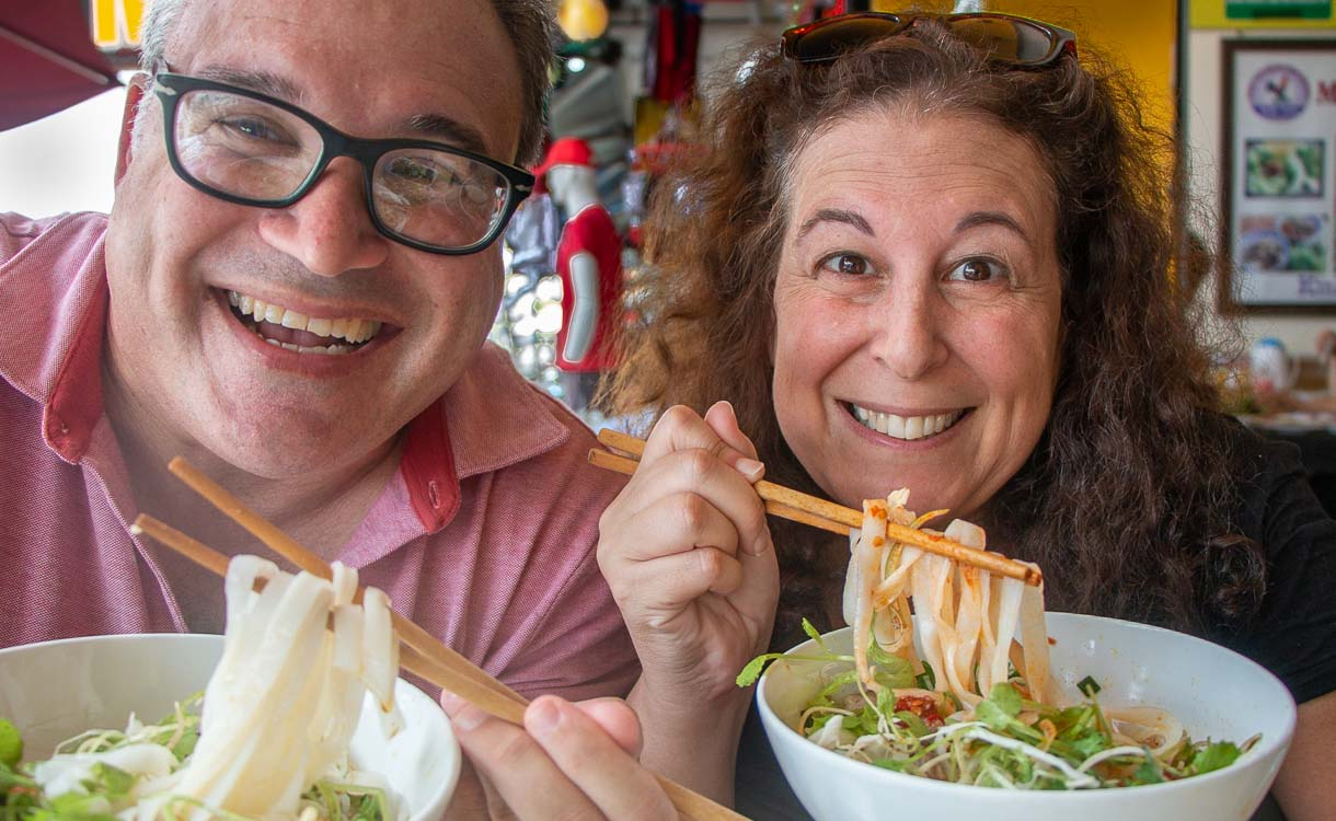 A happy couple taking a selfie, with the man on the left wearing glasses and the woman with curly hair on the right. Both are grinning widely, each holding a white bowl filled with a noodle dish and chopsticks.

