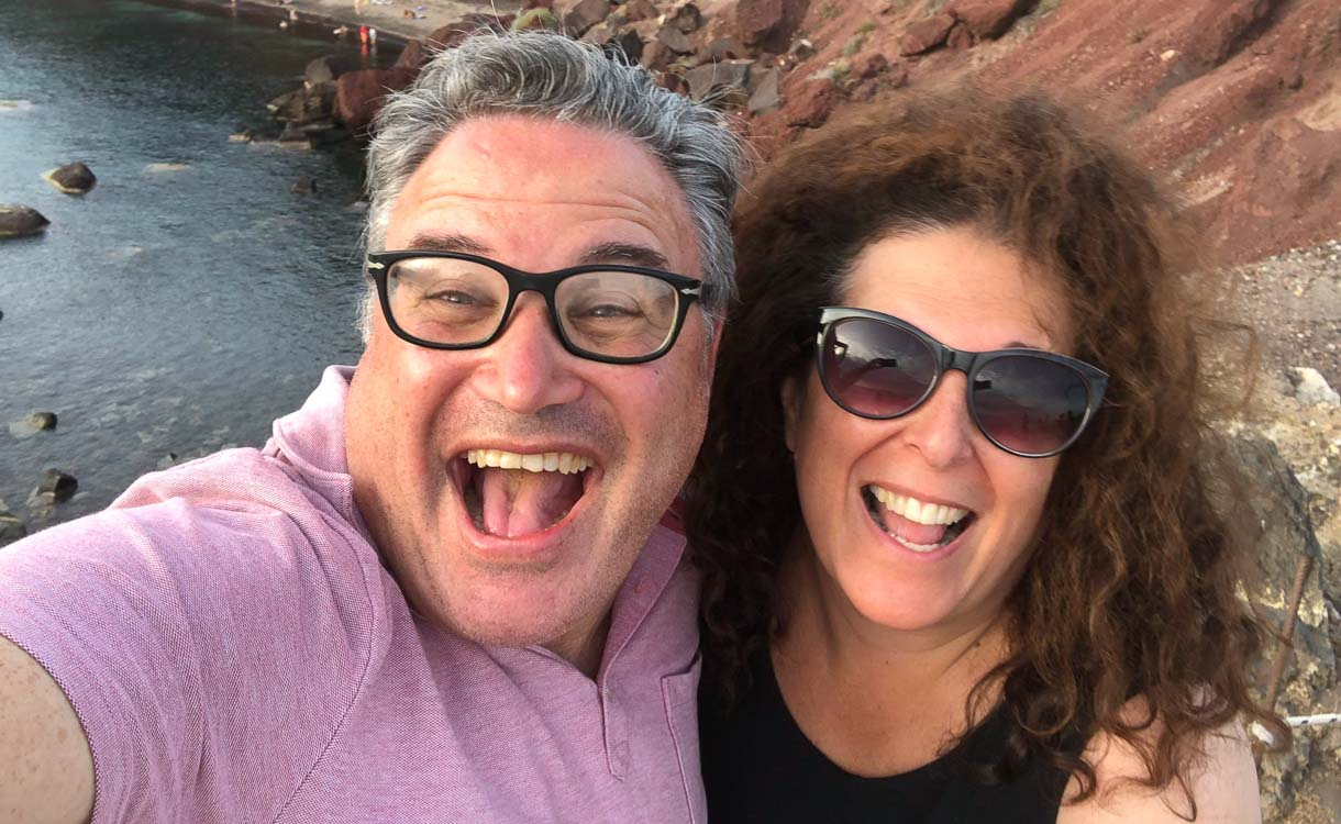 A couple in a high-angle selfie, with brown cliffs and water, likely a lake, behind them. The woman is wearing sunglasses and both are smiling broadly, suggesting an outdoor adventure.
