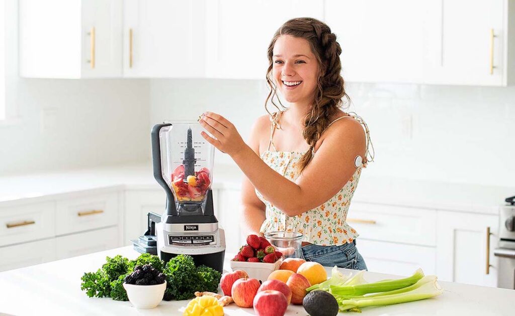 A young woman with braided hair stands in a bright kitchen, smiling as she adds fruits into a blender. The countertop is filled with a variety of fresh produce including strawberries, apples, oranges, celery, and leafy greens. The blender has the brand name 'NINJA' on it. The kitchen features white cabinetry in the background.