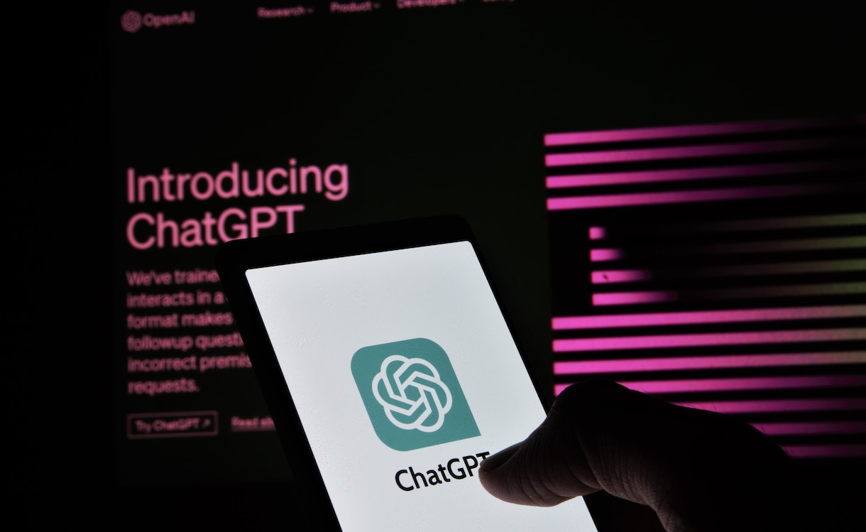 A hand holding a smartphone displays the ChatGPT logo, with the OpenAI website in the background highlighting the introduction of ChatGPT. The website has pink text and graphics on a dark background.
